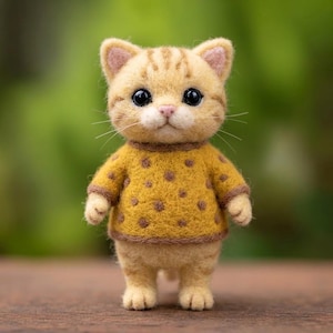 以下が含まれることがあります： 大きな黒い目とピンクの鼻を持つ、ニードルフェルトで作られたクリーム色と茶色のトラ猫のフィギュア。猫は茶色の水玉模様と、襟、袖、裾に茶色の縁取りが施されたマスタードイエローのセーターを着ています。