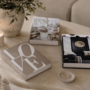 Puede incluir: Tres libros de tapa dura con cubiertas decorativas, una vela beige acanalada en un recipiente de vidrio y un pequeño plato de cerámica con tres anillos dorados. Los libros incluyen los títulos "There is always LOVE," "DECOR," y "SIMPLICITY ETERNAL LUXURY."