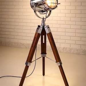May include: A floor lamp featuring a polished chrome spotlight head with a protective metal cage, mounted on a dark brown wooden tripod stand. The lamp includes silver-toned metal hardware at the joints and a black power cord extending downwards.