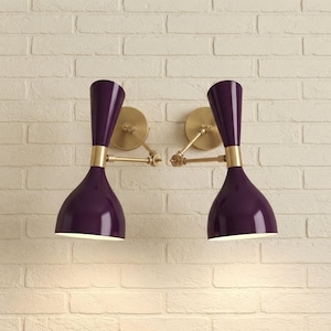 Puede incluir: Dos lámparas de pared con pantallas en forma de reloj de arena de color morado intenso y brazos de metal en tono latón. Cada lámpara tiene una placa circular y un brazo articulado ajustable que conecta la pantalla con la base.