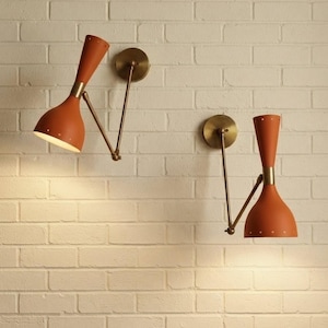 Puede incluir: Dos apliques de pared con pantallas en forma de reloj de arena de color naranja quemado mate y pequeñas perforaciones en los bordes. Cada lámpara tiene un brazo metálico ajustable en tono latón unido a una placa circular de pared.
