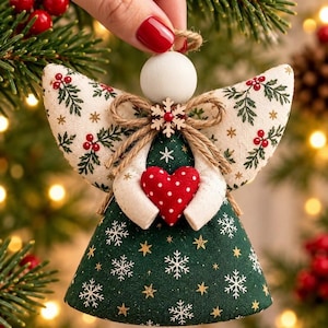 May include: A fabric angel ornament featuring a dark green dress with white snowflakes and gold stars. The wings are cream-colored with a red berry and green leaf pattern. A red heart with white dots is held in the center, accented by a twine bow.