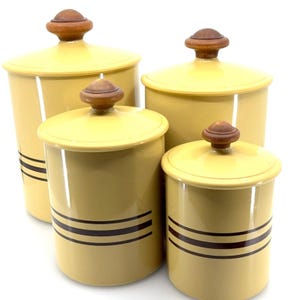 Puede incluir: Un juego de cuatro botes cilíndricos con acabado en amarillo pálido, cada uno con tres franjas horizontales de color marrón oscuro en la base y una tapa a juego rematada con un pomo redondo de madera oscura para facilitar su apertura.