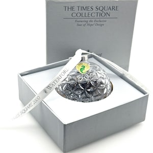 May include: A clear crystal ornament with a faceted star pattern inside a grey box labelled The Times Square Collection. A white ribbon printed with Waterford and Times Square 2000 rests across the ornament, which features a green seahorse sticker.