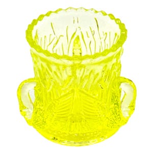 Op de afbeelding: Een felgele, doorschijnende glazen houder met een geschulpte rand en twee handvatten aan de zijkanten. Het buitenoppervlak is versierd met een gegoten, gestructureerd patroon van verticale lijnen en waaiervormige motieven in reliëf.