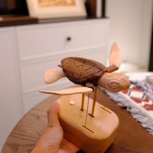 May include: A wooden sea turtle figurine with a dark brown shell and light tan head and flippers. The turtle is mounted on a light-colored wooden base with thin metal rods connecting the flippers and body to the base, allowing for movement.