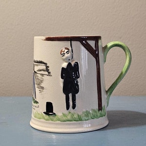 May include: A white ceramic mug with a green handle. The mug has a painted scene of a man hanging from a wooden structure. The scene is set against a backdrop of a waterfall. The mug also has a black hat and the text "Niagara Falls, Canada" painted on it.