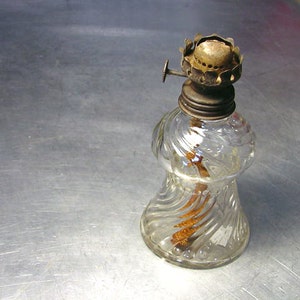 May include: A clear glass oil lamp with a metal wick holder and a decorative swirl pattern. The lamp has a metal cap and a small knob on the side.