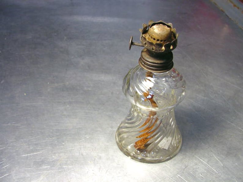 May include: A clear glass oil lamp with a metal wick holder and a decorative swirl pattern. The lamp has a metal cap and a small knob on the side.