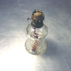May include: A clear glass oil lamp with a swirl design. The lamp has a metal top with a wick and a small metal knob.