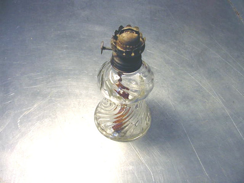 May include: A clear glass oil lamp with a swirl design. The lamp has a metal top with a wick and a small metal knob.