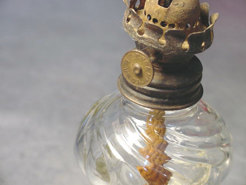 May include: A vintage glass oil lamp with a clear glass reservoir and a brass wick holder. The lamp has a decorative swirl pattern on the glass and a brass cap with a metal knob.