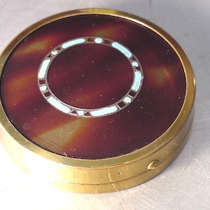 May include: A round, brass compact with a tortoiseshell-like design. The compact has a white and blue enamel ring around the center.