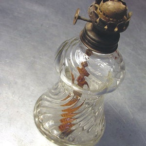 May include: A clear glass oil lamp with a swirl design and a brass top. The lamp has a wick inside and is sitting on a silver surface.