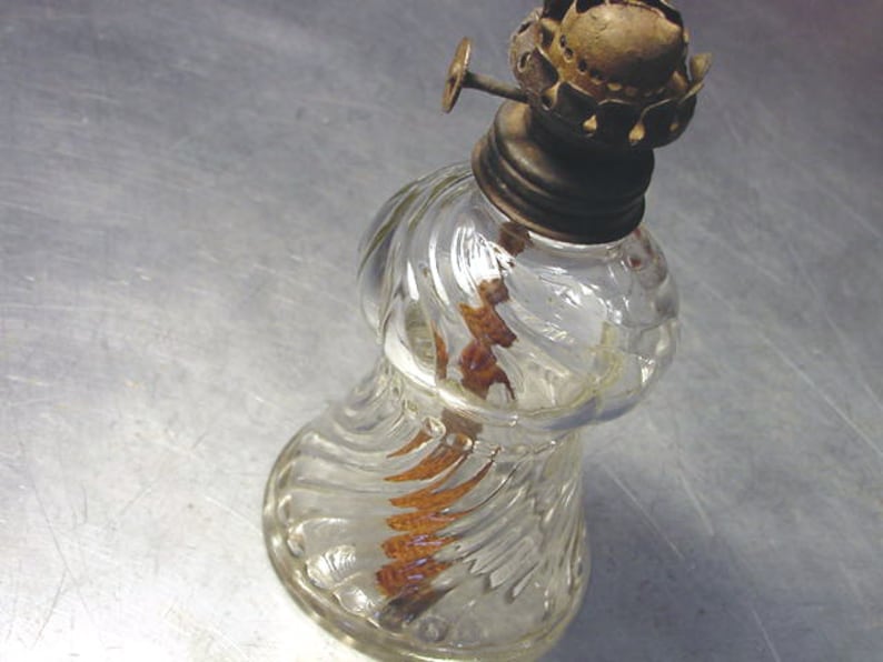 May include: A clear glass oil lamp with a swirl design and a brass top. The lamp has a wick inside and is sitting on a silver surface.