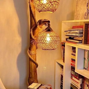 Puede incluir: Una lámpara de pie construida con una pieza vertical de madera flotante natural con dos pantallas esféricas hechas de cuerda beige tejida. La lámpara cuenta con un pequeño estante de madera integrado y una cuerda gruesa alrededor del poste.