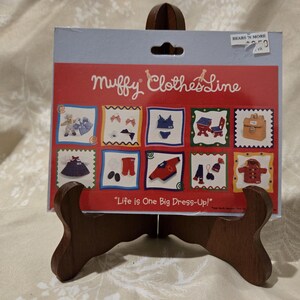 May include: A Muffy Clothes Line product display card featuring the text "Muffy Clothes Line" and "Life is One Big Dress-Up!" with ten illustrated squares showing various doll clothing items, accessories, and furniture on a red background.