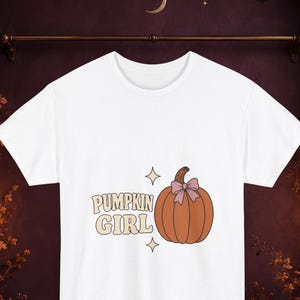 Può includere: Una t-shirt bianca a maniche corte con la grafica di una zucca arancione con un fiocco rosa chiaro sul picciolo. A sinistra della zucca, il testo PUMPKIN GIRL è stampato in un carattere beige in stile retrò, accompagnato da due piccole stelle.