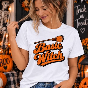 May include: A white short-sleeve t-shirt featuring a graphic print of an orange broomstick above the text Basic Witch in bold orange letters with a black outline. The design has a distressed texture and is centered on the chest.