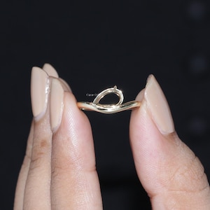 Könnte beinhalten: Ein goldener Ring mit einer zarten, tropfenförmigen Fassung in der Mitte. Die Ringschiene hat eine glatte, polierte Oberfläche und eine sanfte, wellenförmige Kontur. Das Design ist schlicht und raffiniert und zeigt eine minimalistische Metallstruktur.