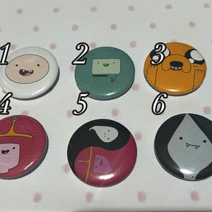 May include: Six circular pin-back badges featuring cartoon character illustrations. Badge 1 is white, 2 is teal, 3 is orange, 4 is pink, 5 is a black and pink yin-yang design, and 6 is a black and grey teardrop shape. Each badge is numbered 1 through 6.