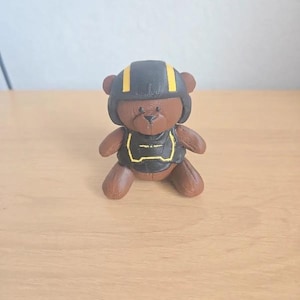 Könnte beinhalten: Eine braune Teddybär-Figur, die einen schwarzen Helm mit gelben Streifen und ein passendes schwarzes Brustteil mit gelber Umrandung trägt. Der Bär hat eine texturierte Oberfläche, kleine Ohren und eine dunkle Nase.