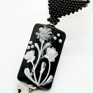 Rectangular lampwork pendant in black with white flowers and a beaded bail. Neckcord included