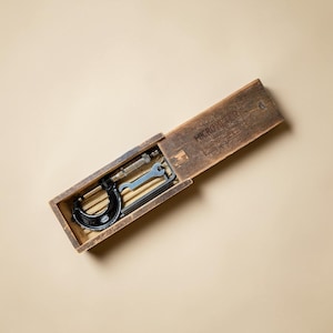 Puede incluir: Un micrómetro de metal negro con un husillo de ajuste plateado y una pequeña llave plateada, guardado dentro de una caja de madera rectangular con tapa deslizante. La palabra MICROMETER está estampada en tinta negra en la tapa de la caja.