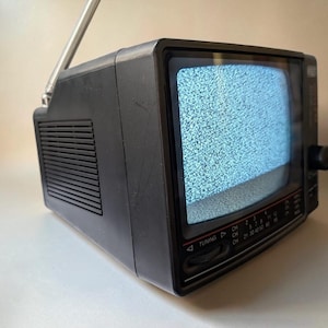 May include: A small black portable television with a static-filled screen, a silver telescoping antenna extended upward, a side speaker grille, and a front-facing tuning dial with channel numbers printed in white text on the lower control panel.