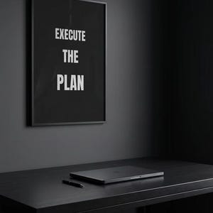 May include: A black framed wall art print featuring the white text EXECUTE THE PLAN in a bold sans-serif font. The artwork is displayed on a dark grey wall above a black desk that holds a closed laptop and a black pen, creating a monochromatic look.