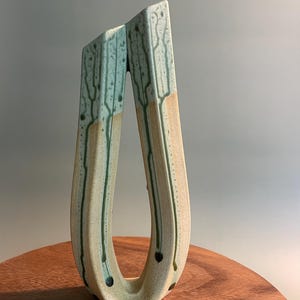 May include: A ceramic vase with a unique, open design. The vase features a light green and tan color scheme with dark green accents. The form is open, with two vertical sides connected at the base, creating an abstract, artistic shape. The vase is placed on a wooden surface.