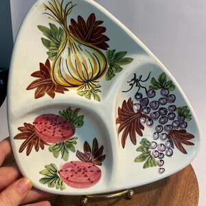 May include: A white, triangular divided serving dish with hand-painted fruit and vegetable designs. The dish features a yellow onion, red fruit, and purple grapes. The dish is on a wooden serving board with a gold-colored metal stand.
