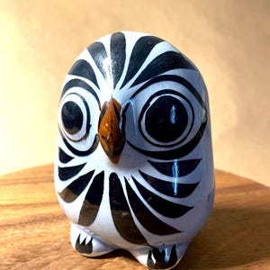 May include: A ceramic owl figurine with a white base, black markings, and large black eyes. The owl has a brown beak and is sitting on a wooden surface. The owl's design is a stylized representation of the bird.