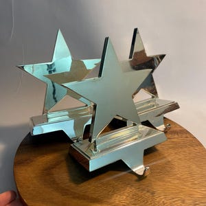 May include: Silver star-shaped stocking holders. Each holder features a star design with a hook for hanging stockings. The holders are displayed on a round wooden surface, creating a festive holiday decoration.