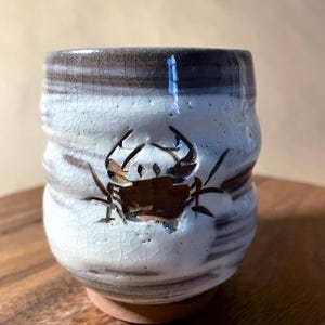 May include: A handmade ceramic cup with a crab design. The cup features a white and brown glaze with a textured, crackled surface. The crab is depicted in brown, adding a rustic, coastal theme.