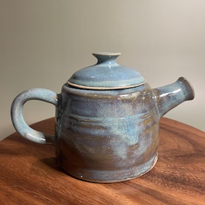 May include: A handmade ceramic teapot with a blue and brown glaze. The teapot has a curved handle, a spout, and a lid with a small knob. The teapot is sitting on a wooden surface.