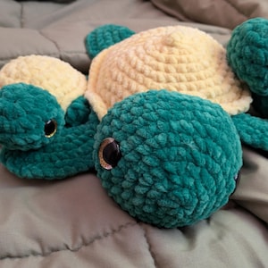 Puede incluir: Un peluche de tortuga grande hecho a ganchillo con un caparazón central de color crema y cabeza y aletas de color verde azulado. Dos tortugas de ganchillo más pequeñas están unidas a los lados, cada una con caparazón crema y cabeza y aletas verde azulado.