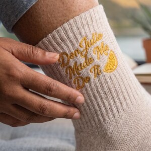 May include: A beige ribbed crew sock featuring gold embroidered text that reads Don Julio Made Me Do It, accompanied by a small embroidered yellow lemon wedge graphic. The sock is made of a knit fabric and has a textured, vertical ribbed pattern.