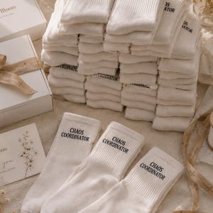 May include: A stack of white ribbed crew socks featuring the black text "CHAOS COORDINATOR" printed on the cuff. Three individual socks are laid out in front of the stack, each displaying the same black text on the ribbed upper section.