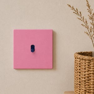 May include: A square light switch plate in a solid matte pink finish featuring a single dark blue toggle switch centered on the face. The switch plate is mounted flush against a plain wall surface, providing a clean and simple aesthetic.