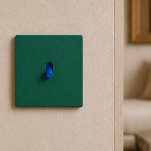 May include: A square light switch plate in a matte dark green finish featuring a single centered toggle switch in a vibrant blue color. The switch plate has a smooth, flat surface and is mounted flush against a light beige textured wall.