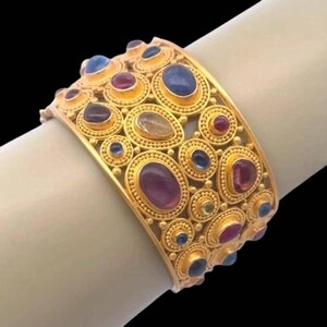 Puede incluir: Una pulsera brazalete ancha de oro con una superficie texturizada y detalles de metal granulado. La pulsera está adornada con numerosas piedras preciosas cabujón ovaladas y redondas en tonos azul, morado, rojo y claro, engastadas en oro.