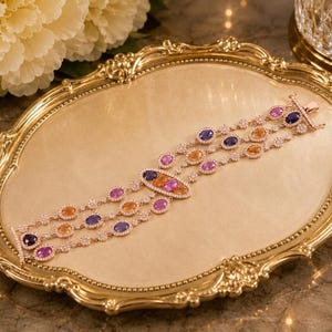 May include: A multi-strand bracelet crafted in rose gold, featuring three parallel chains adorned with oval-cut sapphires in shades of pink, orange, and deep blue. Each gemstone is surrounded by a halo of small, clear, sparkling white stones.
