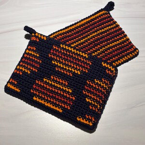 May include: Two handmade crochet potholders. One is navy blue with orange and yellow heart shapes. The other is navy blue with orange and yellow stripes. Both have a loop for hanging.