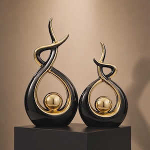 Op de afbeelding: Twee abstracte sculpturen met een glanzende zwarte buitenkant en een reflecterende gouden binnenkant. Elk stuk heeft een gebogen, vlamachtig silhouet dat een centrale, gladde gouden bol omlijst. De sculpturen staan naast elkaar.