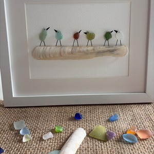 Può includere: Un'opera d'arte da parete incorniciata raffigurante sei uccelli realizzati con vetro marino colorato appollaiati su una lunga conchiglia chiara. Gli uccelli hanno zampe e becchi in filo metallico nero. Sotto la cornice, vari pezzi di vetro marino.