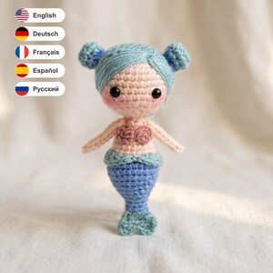 May include: A small crocheted mermaid doll featuring light blue yarn hair styled in two buns, a pale peach-coloured face with black bead eyes and pink cheeks, a light pink shell-shaped top, and a textured blue tail with a light blue fin.