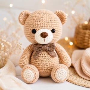 Puede incluir: Un oso de peluche de ganchillo color canela claro con un hocico blanco, una nariz marrón oscuro y ojos de plástico negro. El oso lleva una pajarita de ganchillo marrón oscuro alrededor del cuello y parches blancos en las plantas de los pies.