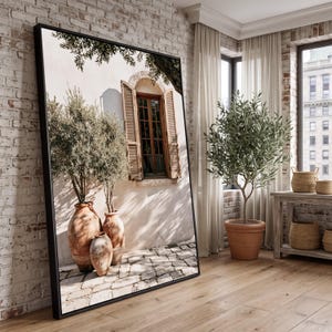 Op de afbeelding: Een groot rechthoekig ingelijst wandkunstwerk met een zonnige mediterrane scène met drie terracotta potten, olijfbomen en een raam met houten luiken op een lichte muur, geplaatst tegen een witte bakstenen muur in een kamer.