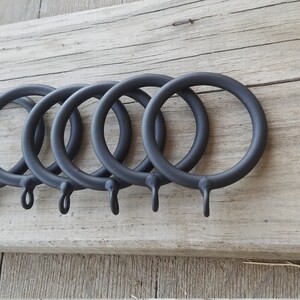 May include: Six black metal curtain rings with a hook on each ring. The rings are arranged in a row on a wooden surface.